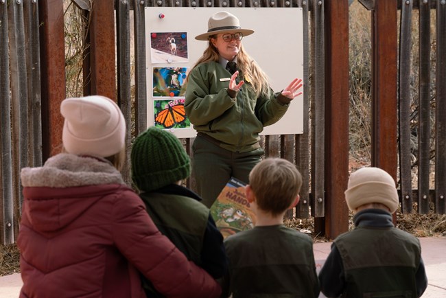 A ranger in a green coat stands in front of a seated family in hats and coats.