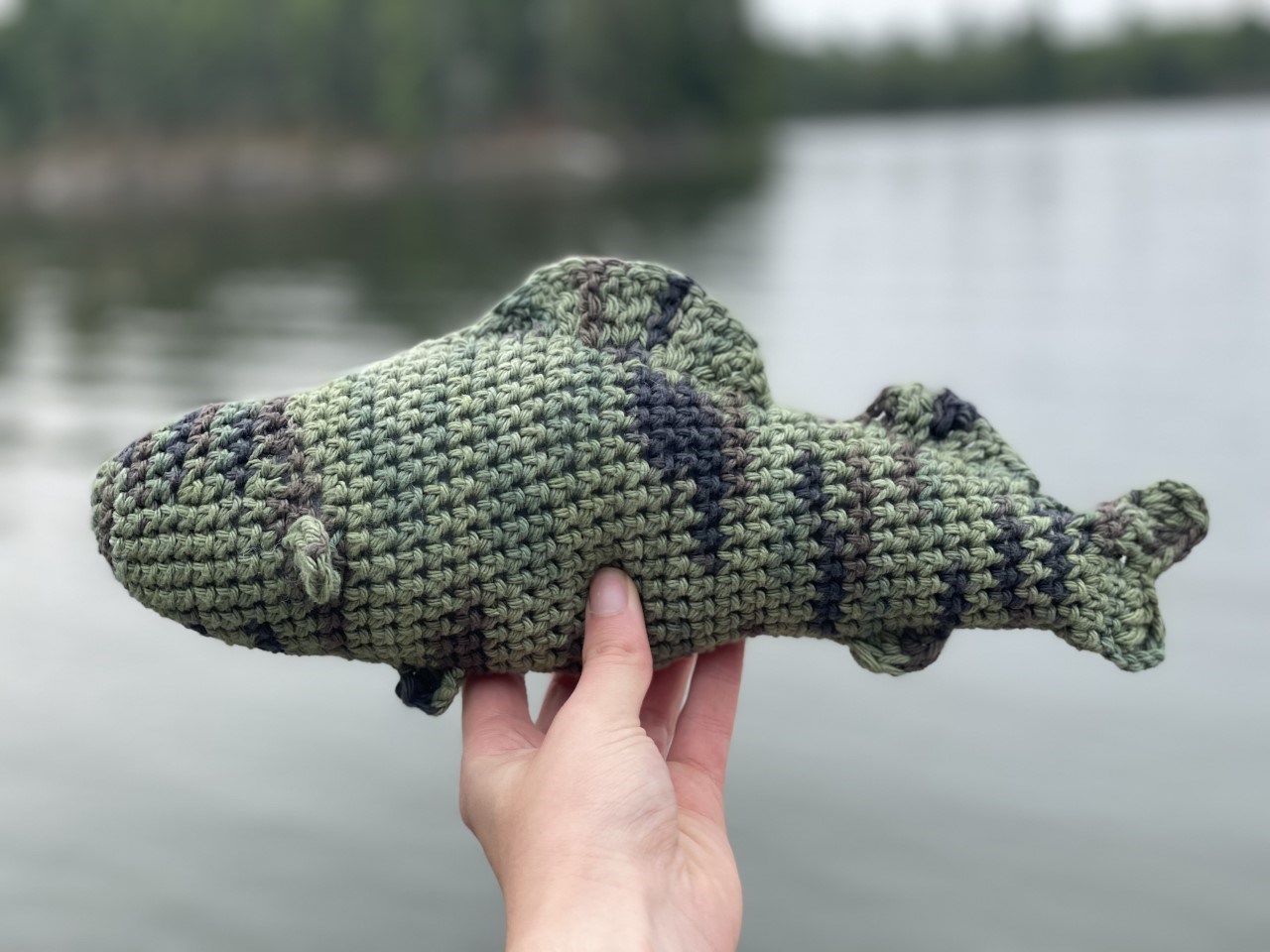 Crochet fish A crocheted fishing being held by a hand in front of a lake.