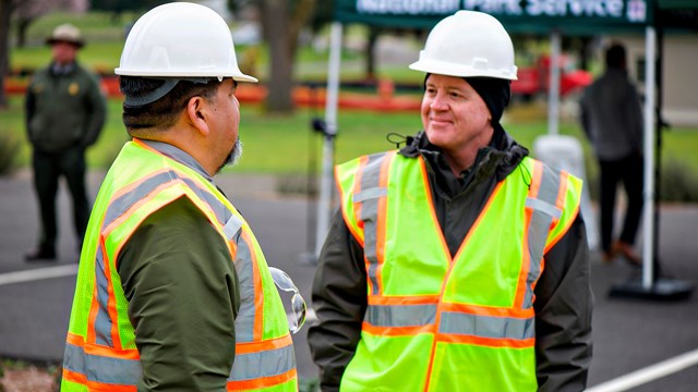 Two men wearing reflective vests and hard hats talk outdoors.