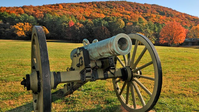 Vintage cannon displayed on grass field in front of distant hill covered in fall colors.