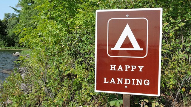 A brown sign with a tent symbol labeled "Happy Landing" stands near some shrubs by a lake.