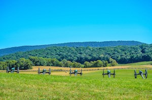 a row of five cannons sit in a field pointed toward a distant tree-covered ridge; large hay bails and worm fence are in between the cannons and the ridge